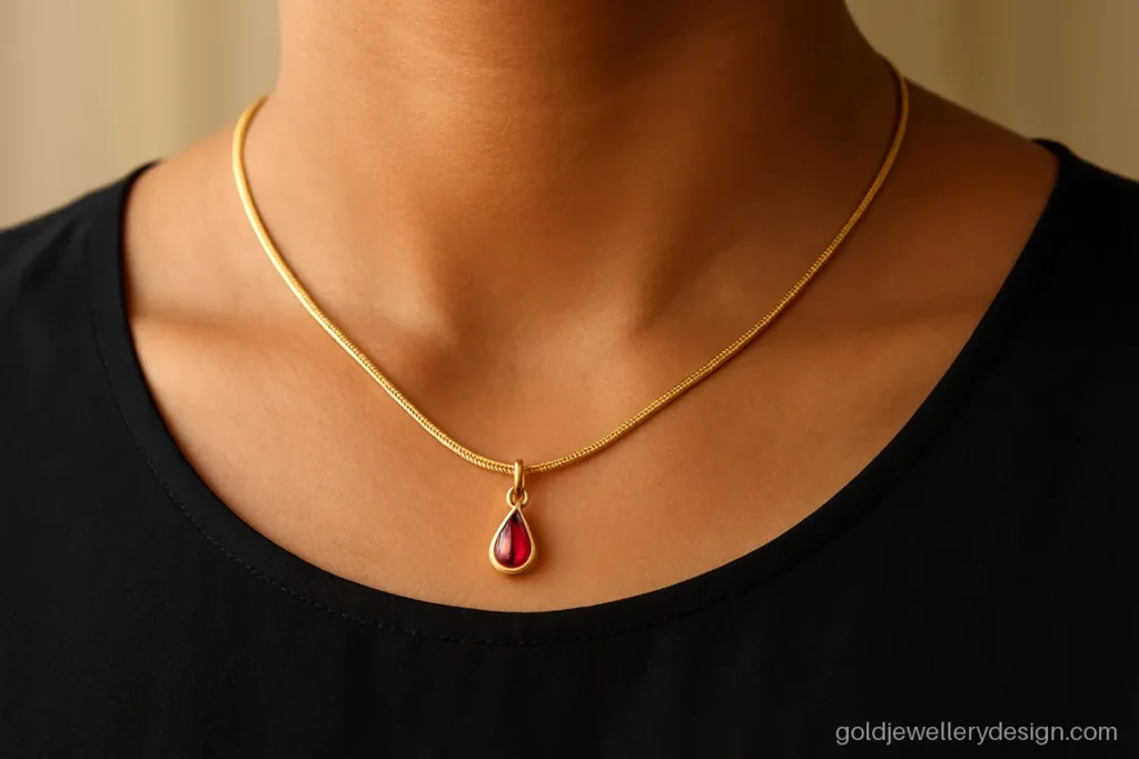Snake link gold chain with a small ruby drop pendant worn on the collarbone